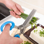 Scissors cutting green onions with a hand holding them above a tray of food.