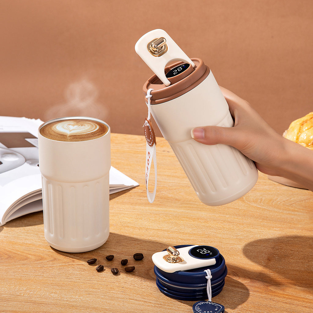 Hand holding a white travel mug with a coffee cup on a wooden table.