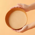 Hand holding a round air fryer paper tray against an orange background.