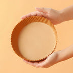 Hand holding a round air fryer paper tray against an orange background.